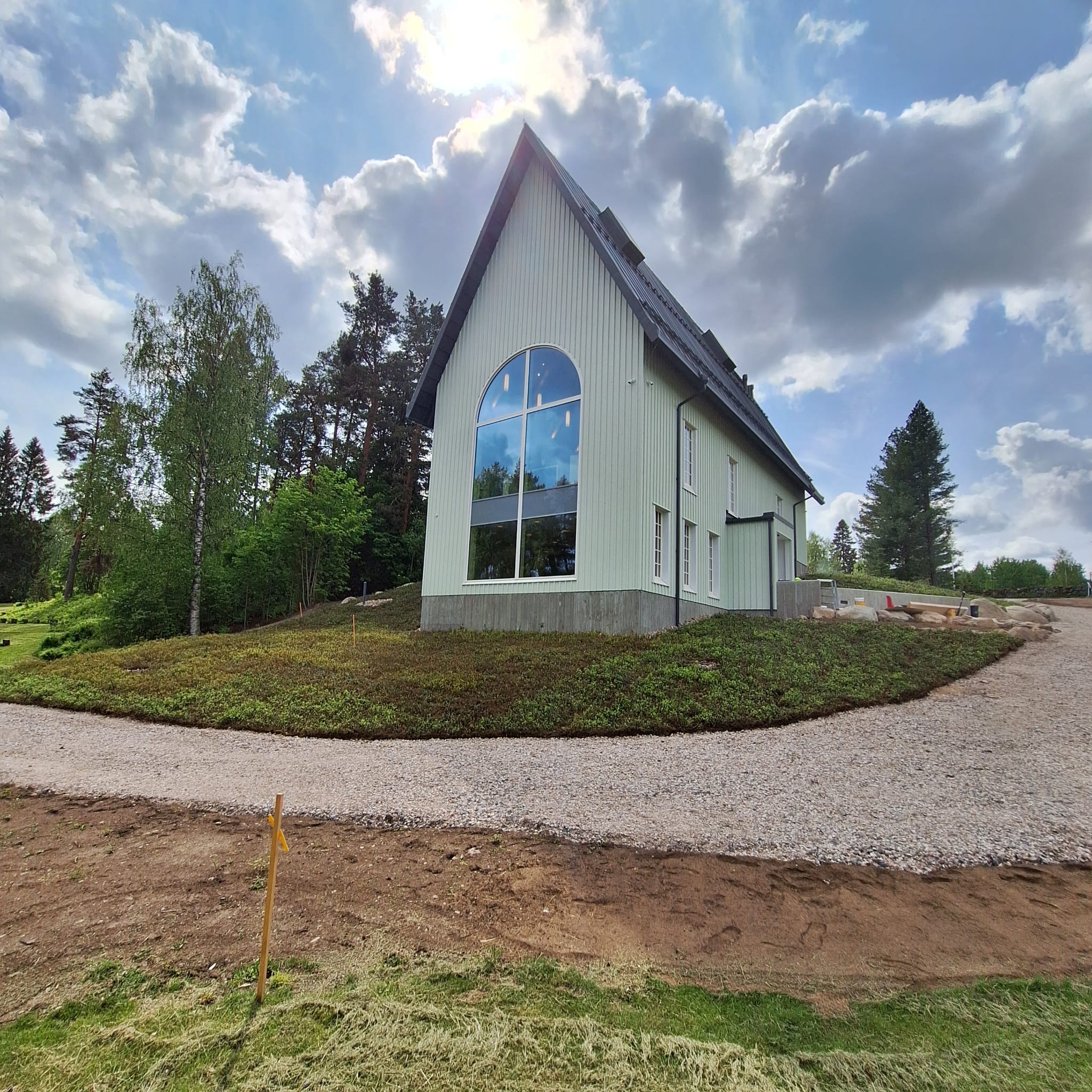 Ulkokuva kirkon päädystä ja sivusta. Maahan asennettu kunta, kivituhkatie edessä. Pilvinen taivas.
