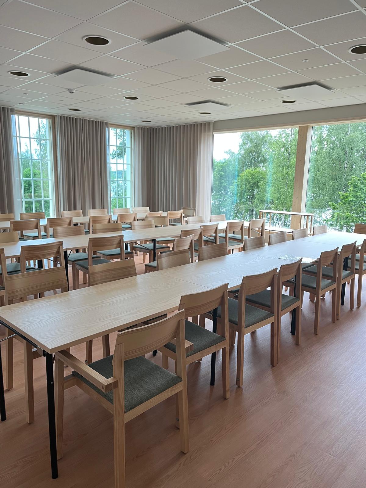 Kuvassa seurakuntasali. Kolme pöytää, joissa on tuolit ympärillä. Taustalla ikkunasta näkyy metsää.